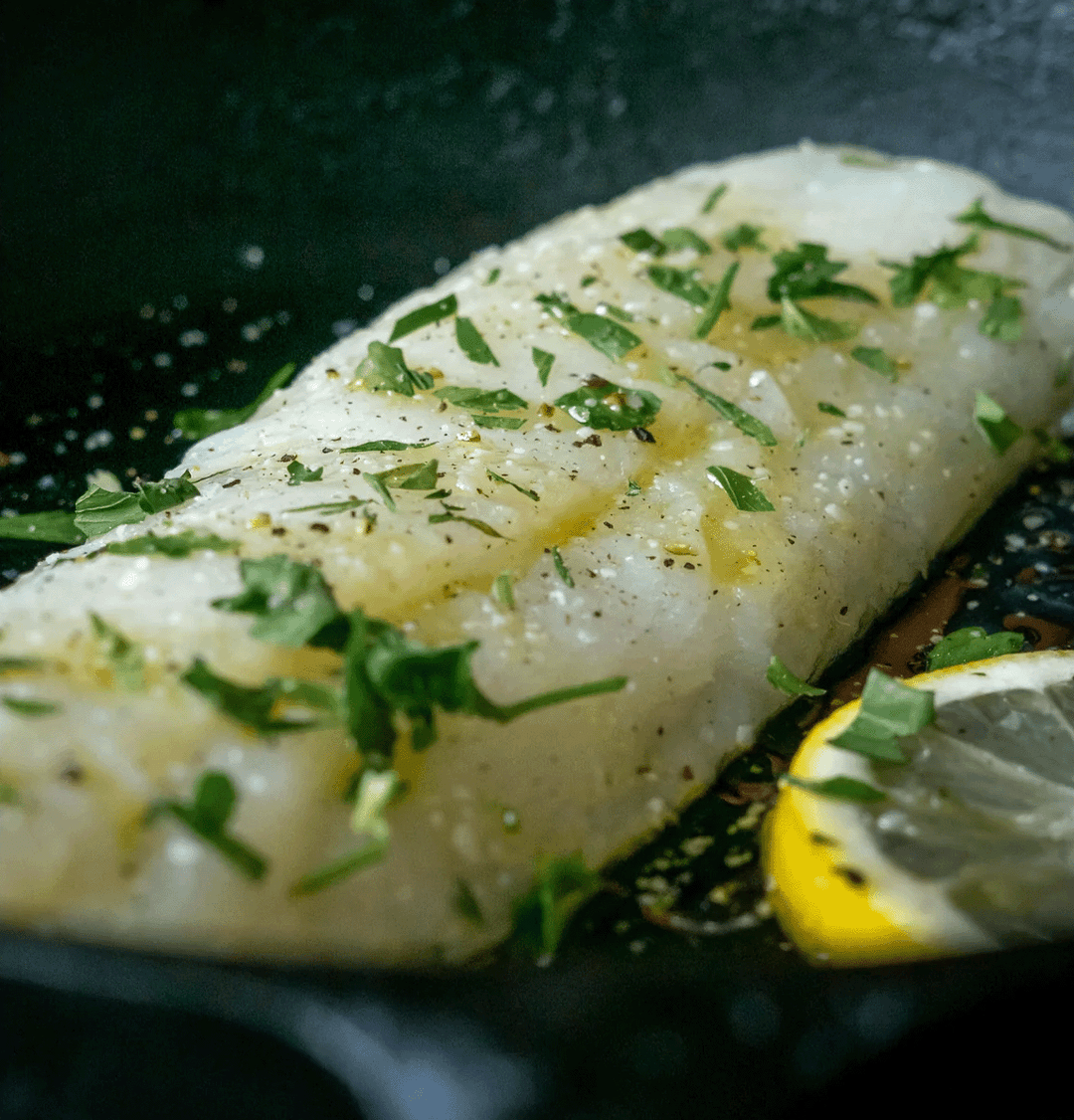 The image shows a piece of Alaskan Pacific cod being cooked in a pan. The fillet is lightly seasoned with salt, pepper, and drizzled with olive oil, and it is garnished with fresh chopped herbs, possibly parsley. A lemon slice is placed beside the fillet, adding a bright accent to the dish. The fillet has a delicate, moist texture, and the herbs and seasonings are visible on its surface. The pan’s dark background contrasts with the light color of the fish, emphasizing its freshness and preparation.