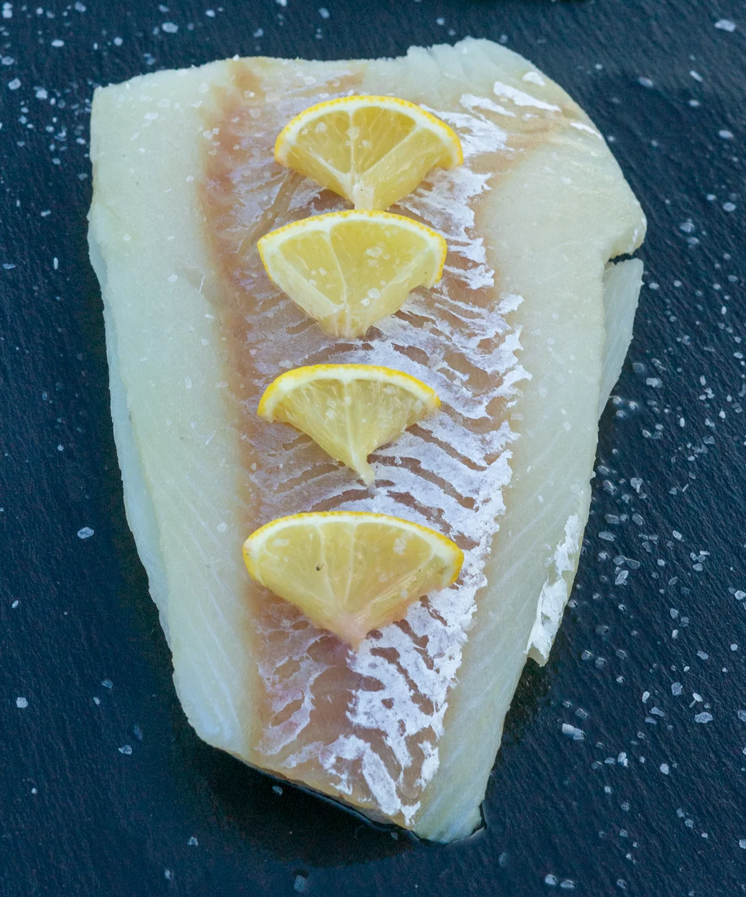 The image displays a raw fillet of Alaskan Pacific cod placed on a dark slate surface. The fillet has a translucent, slightly glossy texture with a natural gradient from white to light pink. Four small lemon wedges are neatly arranged in a row along the center of the fillet, adding a pop of color. Coarse sea salt crystals are sprinkled around the fillet, enhancing its fresh and natural presentation. The overall setting highlights the simplicity and freshness of the seafood.